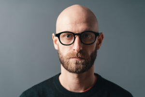 a bald man with a beard and glasses stares into the camera